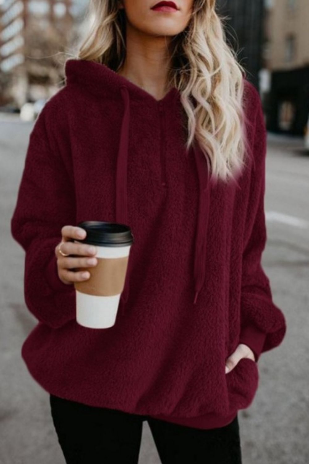 Woman wearing a burgundy quarter-zip drawstring teddy hoodie, holding a coffee cup, standing on a city street.