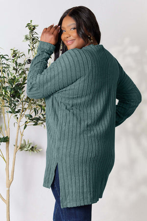 Curvy model showcasing a ribbed, long sleeve, high-low slit top in green, styled with plant decor.