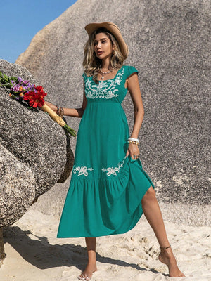 Model wearing an embroidered square neck cap sleeve dress in teal, standing on the beach with flowers.