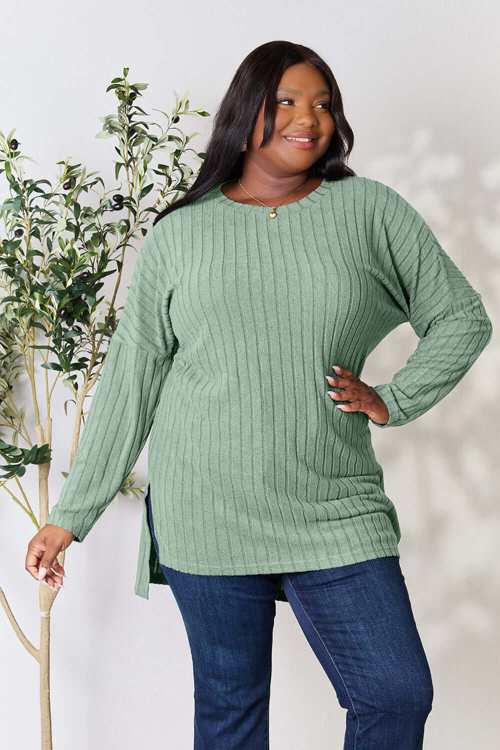 Plus size model wearing a ribbed green long sleeve top with slit detail, paired with jeans, in a chic indoor setting.