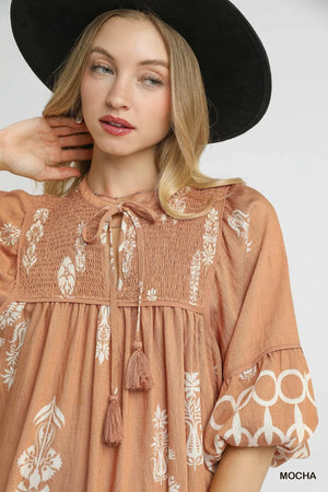 Model wearing Umgee Boarder Print Tassel Tie Neck Mini Dress in mocha color, showcasing bohemian style and vibrant border print.