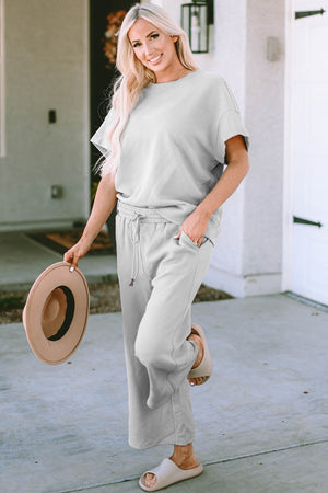 Model wearing the Double Take Full Size Two-Piece Short Sleeve Top and Pants Set in light gray with a hat and sandals.