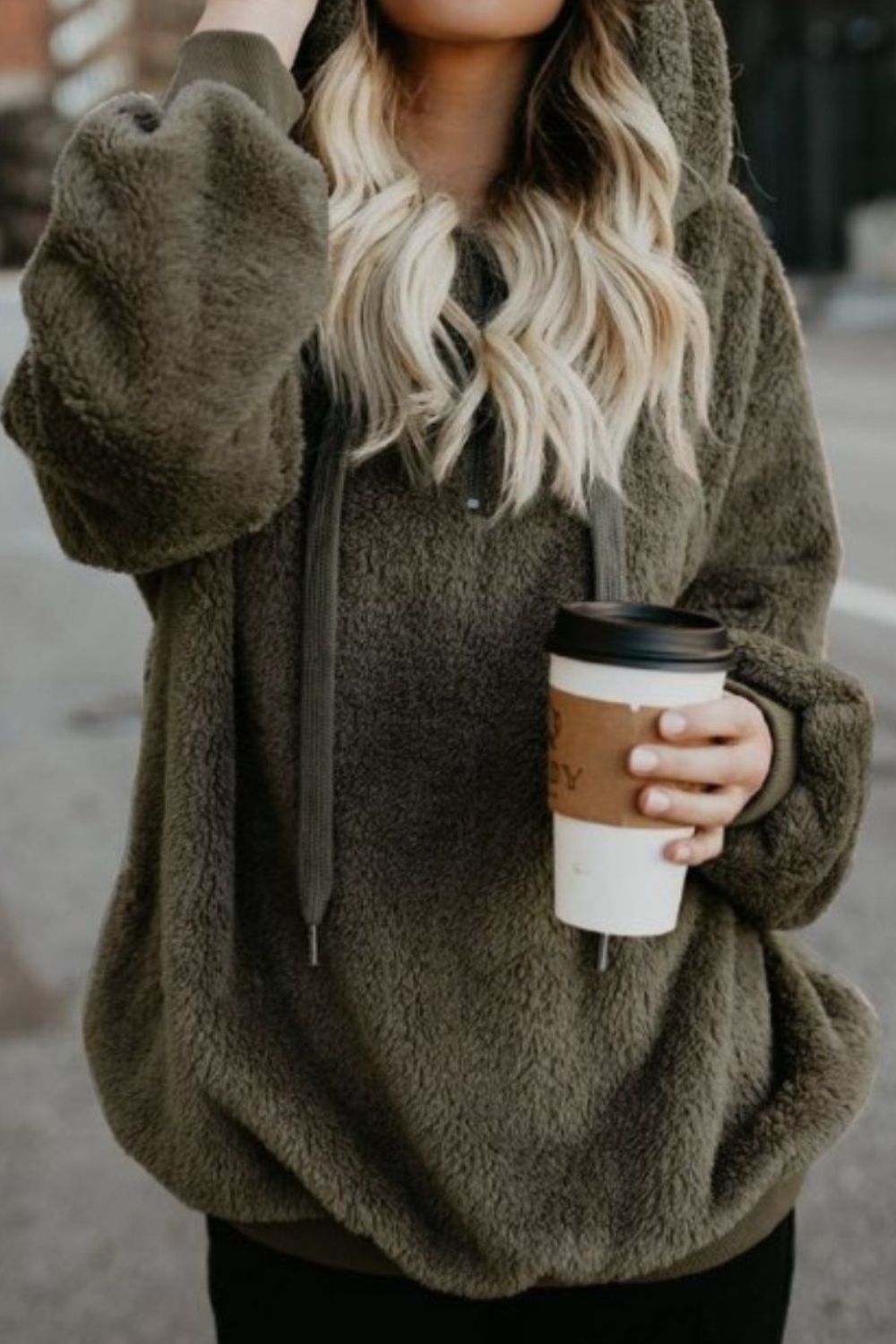 Woman wearing olive green quarter-zip drawstring teddy hoodie, holding a coffee cup, outdoors.