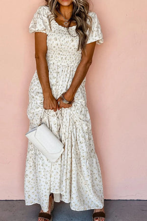 Model wearing a floral smocked short puff sleeve maxi dress, holding a white handbag against a pink background.