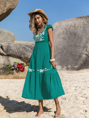 Model wearing an embroidered square neck cap sleeve dress in teal at the beach, holding flowers and wearing a hat.