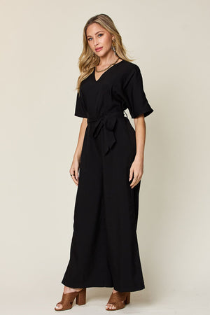 Woman in a black full size V-neck tie front jumpsuit, featuring a slit and stylish bell sleeves.