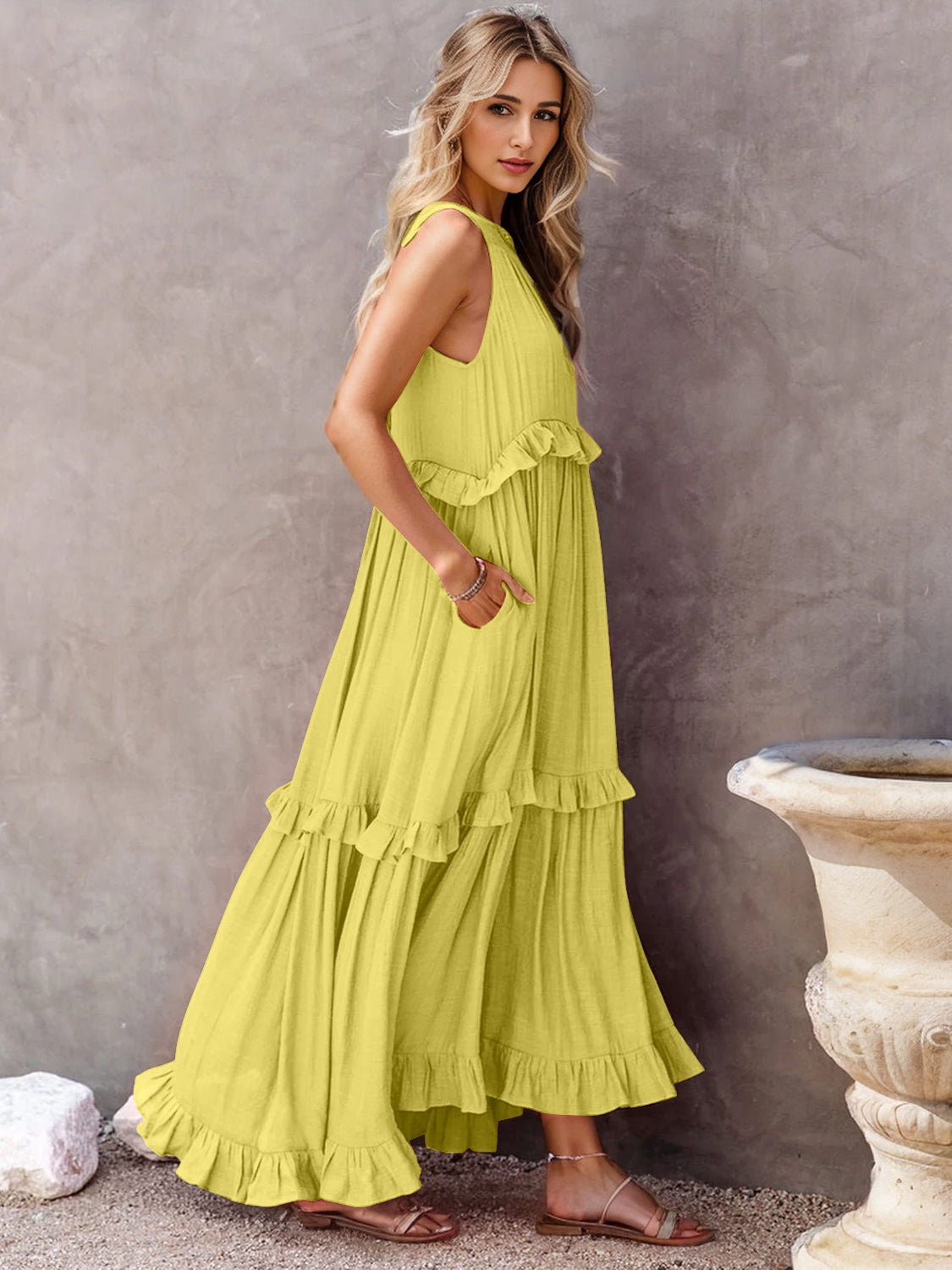 Model wearing a vibrant yellow ruffled sleeveless tiered maxi dress with pockets against a textured background.