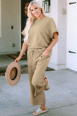 Model wearing Double Take Full Size Texture Short Sleeve Top and Pants Set, tan color, outdoors, holding a hat.