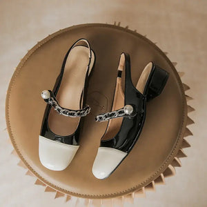 Stylish contrast round toe block heel Mary Jane shoes in black and cream, featuring chain accents, displayed on a round leather surface.
