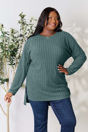 Plus-size model wearing a green ribbed long sleeve top with a slit, paired with denim, against a plant background.