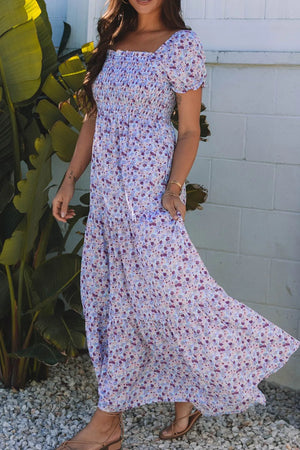 Model wearing a tiered floral shirred high waist maxi dress in light blue, surrounded by tropical plants.