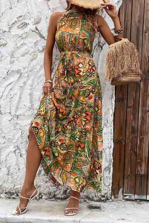 Model wearing a tied slit floral sleeveless dress with vibrant patterns, paired with a straw hat and bag.