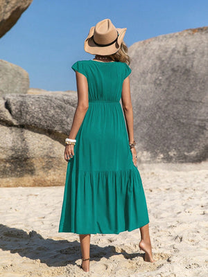 Back view of a woman in a green embroidered square neck cap sleeve dress on the beach, wearing a sun hat.