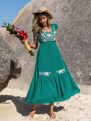 Woman wearing an embroidered square neck cap sleeve dress, holding flowers on a beach with rocks in the background.