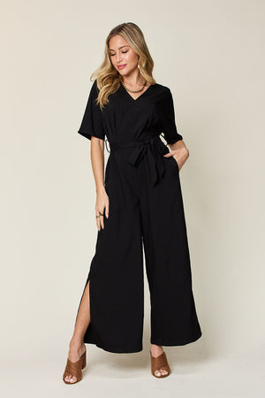 Model wearing a full-size black V-neck tie front jumpsuit with a slit, styled for a chic look.