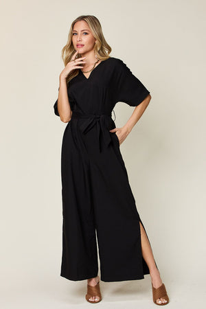 Model wearing a full size black V-neck tie front jumpsuit with slit details and loose sleeves.