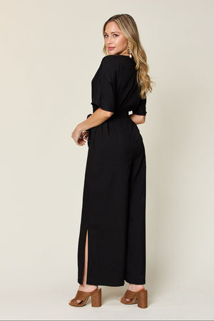Woman modeling a black full size V-neck tie front jumpsuit with slit details, perfect for casual or semi-formal occasions.