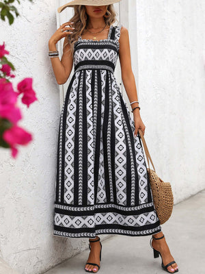 Woman in a black and white printed square neck wide strap cami dress, posing outdoors with a straw bag.