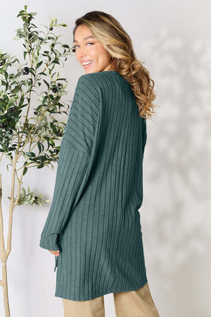 Model wearing a ribbed turquoise long sleeve top, smiling with hair down, surrounded by greenery.