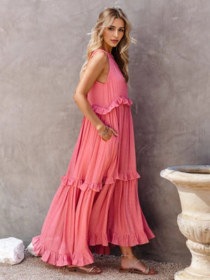 Ruffled sleeveless tiered maxi dress in pink with pockets, styled against a textured wall.