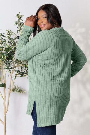 Plus-size model wearing a ribbed green long sleeve top with side slits, standing next to a plant.