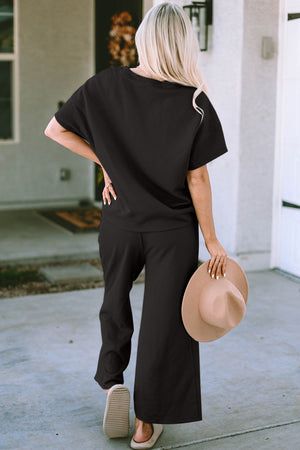 Back view of a woman in a black short sleeve top and pants set, holding a hat, showcasing casual style.