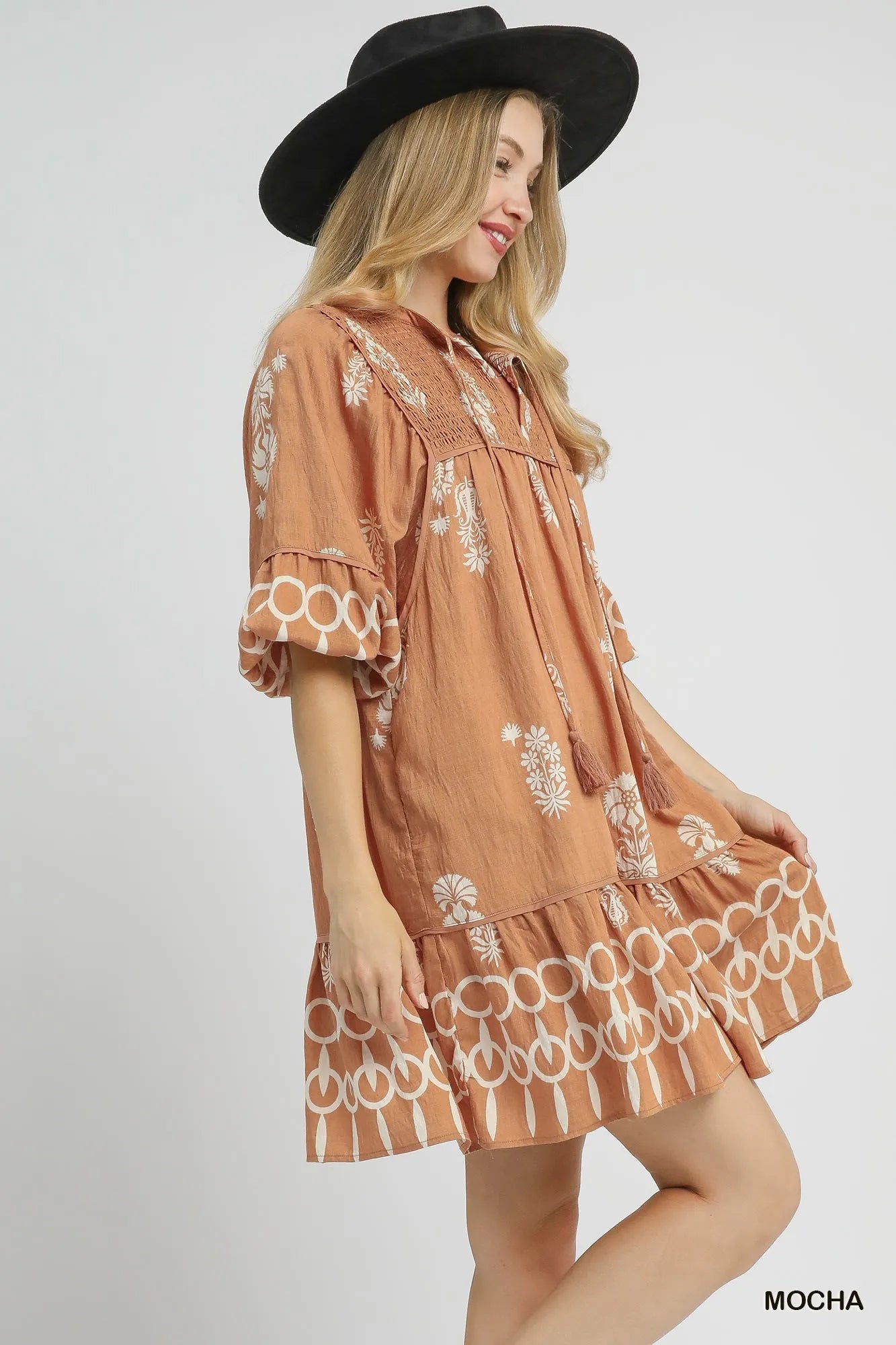 Model wearing a bohemian-inspired brown mini dress with floral prints and ruffled hem, paired with a stylish black hat.