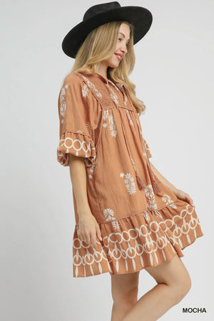 Model wearing a bohemian-inspired brown mini dress with floral prints and ruffled hem, paired with a stylish black hat.