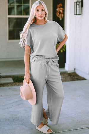 Model wearing the Double Take Full Size Texture Short Sleeve Top and Pants Set in gray, holding a hat, showcasing a casual, stylish look.