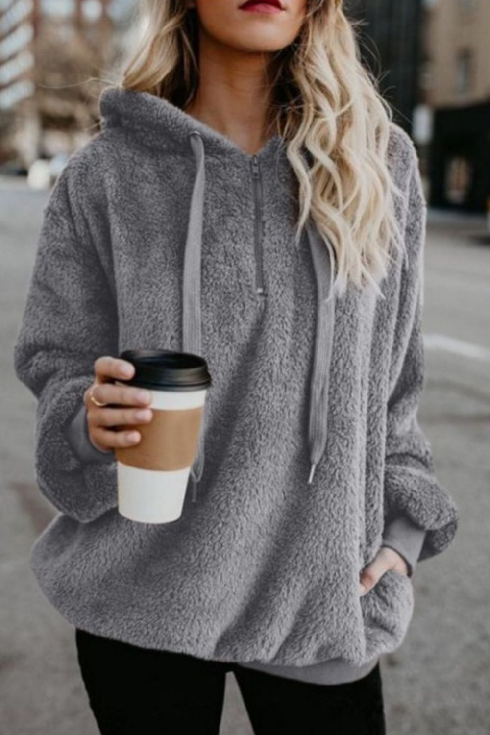 Stylish woman wearing a gray fleece quarter-zip hoodie, holding a coffee cup on a city street.