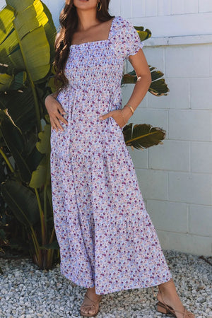 Model wearing a tiered floral shirred high waist maxi dress in purple against a tropical backdrop.