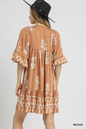 Back view of a woman in a mocha tiered mini dress with ruffled sleeves and floral border print.