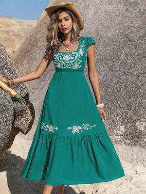 Woman wearing an embroidered square neck cap sleeve dress in teal, showcasing floral designs and a flowy skirt.