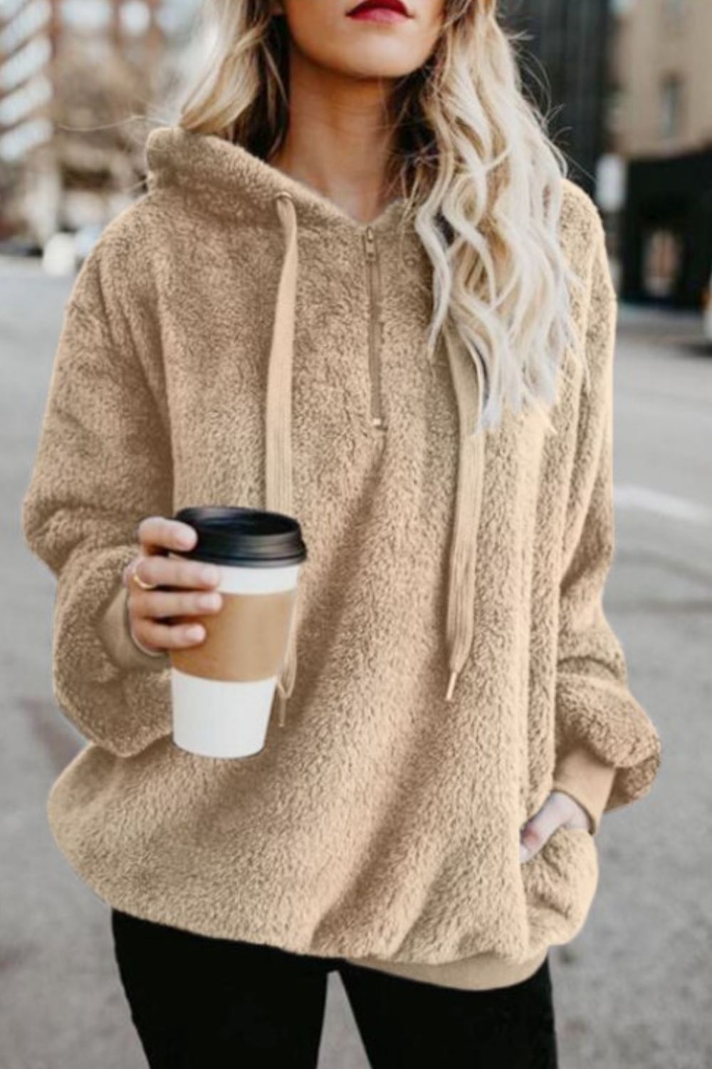 Beige quarter-zip drawstring teddy hoodie with pockets, held by a woman sipping coffee outdoors.