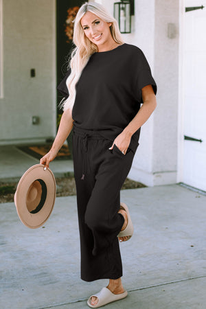 Woman wearing a stylish black short sleeve top and pants set, holding a hat, standing outside.