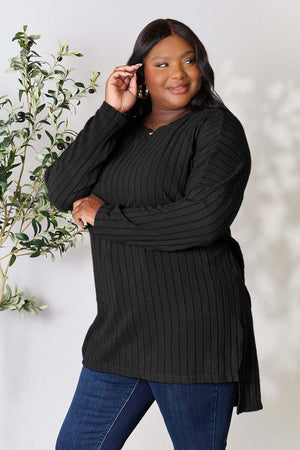 Plus size model wearing Basic Bae ribbed round neck long sleeve slit top in black, styled with jeans and standing by a plant.