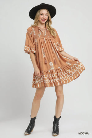 Umgee Boarder Print Tassel Tie Neck Mini Dress in mocha color, featuring a vibrant border print and playful tassels, styled with a hat.