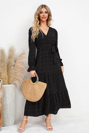 Model wearing a black Swiss Dot Tied Surplice Flounce Sleeve Dress, holding a straw bag, showcasing its stylish design.