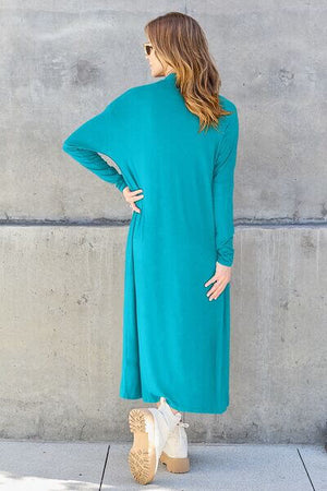 Model wearing a long sleeve open front cover up in teal, showcasing its pocketed design against a textured background.