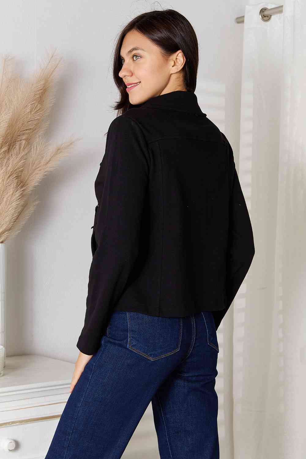Back view of a woman wearing a black full size zip-up jacket with pockets and casual jeans.