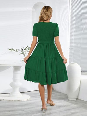 Back view of a woman wearing a green smocked tie front short sleeve tiered dress in a bright room.