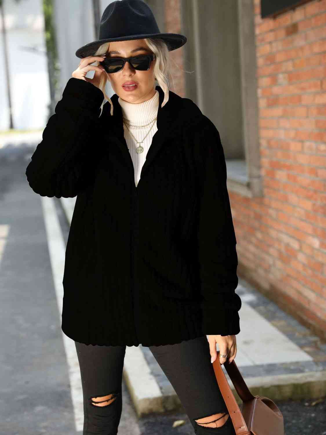 Woman in stylish zip-up collared ribbed jacket, black, paired with sunglasses and a hat, walking outdoors.