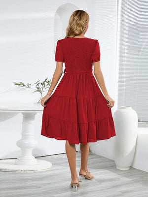 Back view of a red smocked tie front short sleeve tiered dress in a stylish indoor setting.