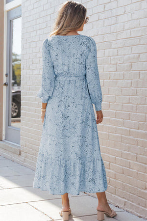 Back view of a woman wearing a printed tie front ruffle hem dress in light blue against a brick wall.