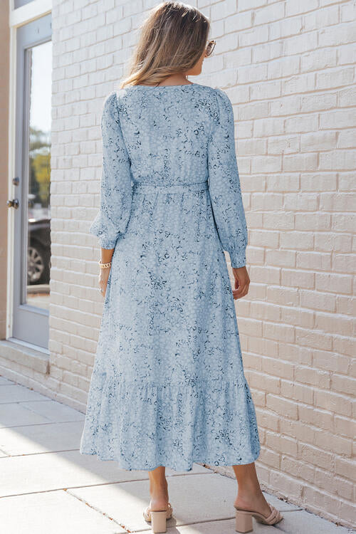 Back view of a woman wearing a printed tie front ruffle hem dress in light blue against a brick wall.