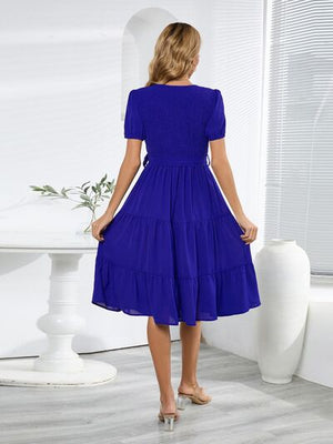 Model wearing a blue smocked tie front short sleeve tiered dress, showcasing the back view in a bright indoor setting.