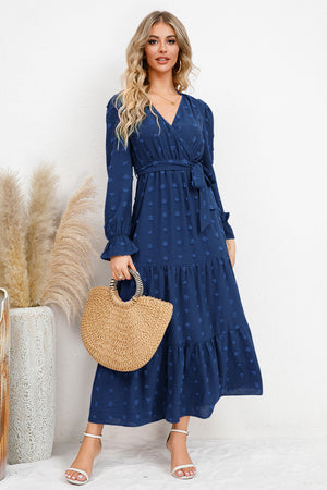 Model wearing a Swiss Dot Tied Surplice Flounce Sleeve Dress in navy, holding a straw bag with a stylish backdrop.