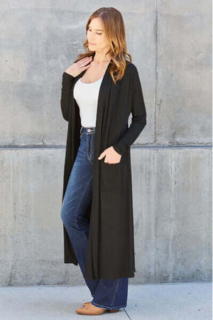 Model wearing a black long sleeve open front cover up with pockets over jeans and a white top.