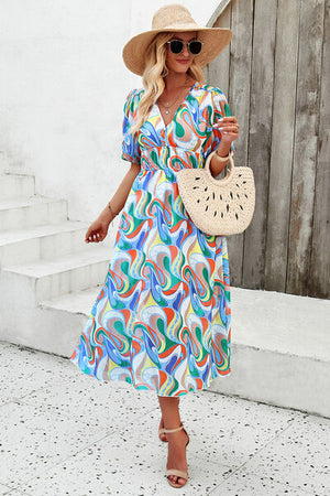 Model wearing a vibrant, smocked printed V-neck short sleeve dress with a straw hat and bag in a sunny outdoor setting.