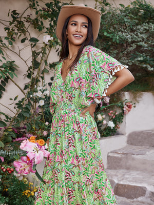 Model wearing a vibrant green floral dress with short sleeves, holding flowers, against a botanical backdrop.